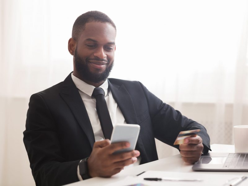 Black manager holding credit card and smartphone, making online purchases at office, copy space
