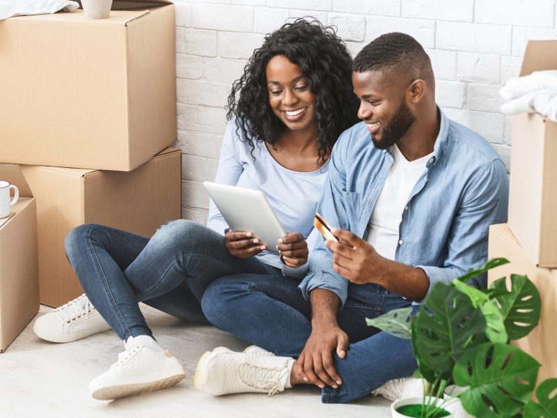 Cheerful african couple purchasing new furniture online, using credit card and digital tablet, just moved in new house, free space